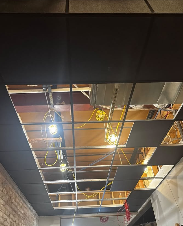 Construction: partially installed black ceiling tiles, exposed framework, and hanging work lights.