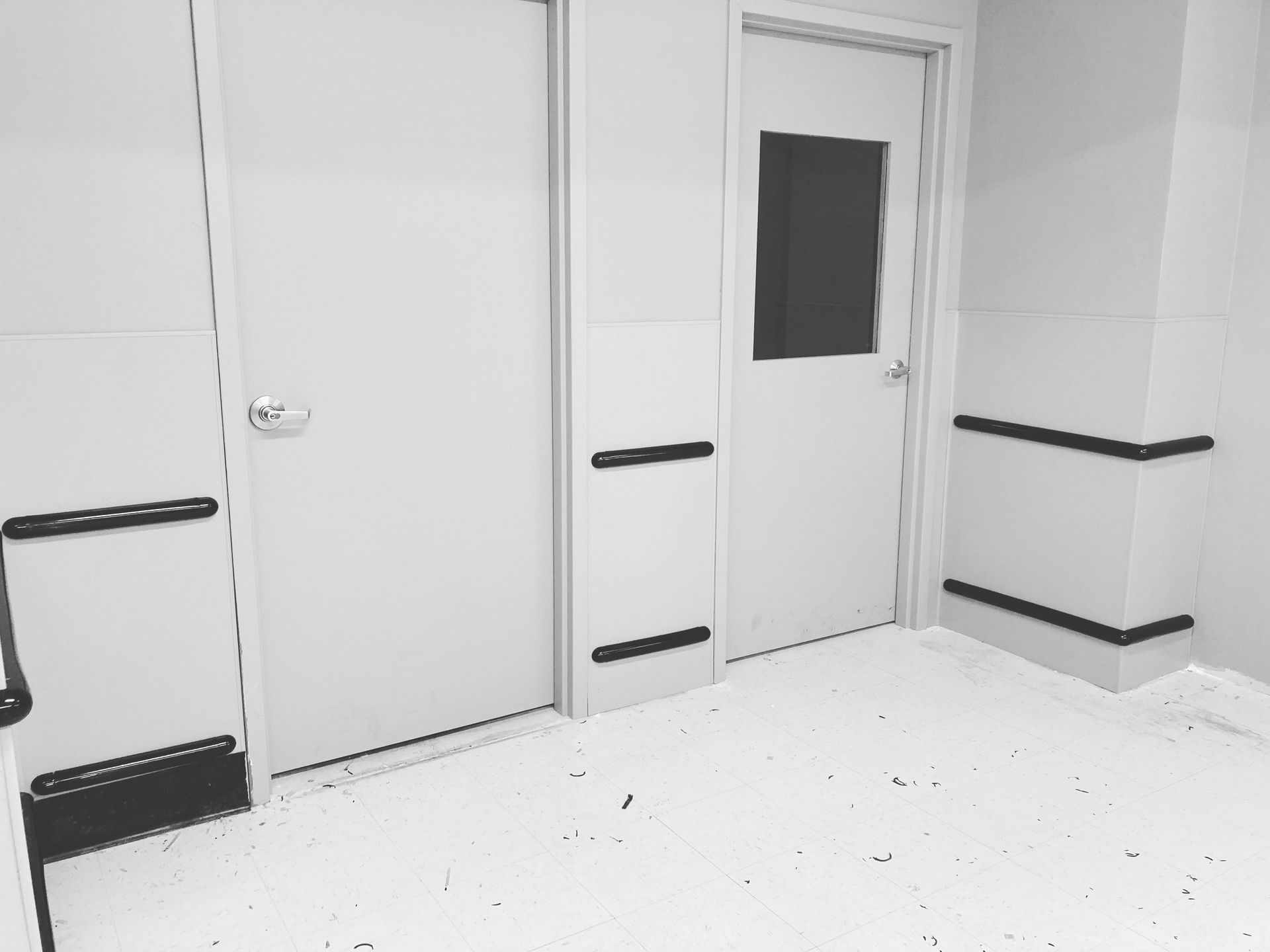 Hallway with white walls, two closed doors, black rectangular bumpers, and a dark window in one door.