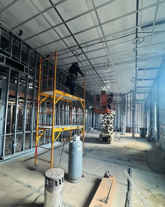 Construction site interior: metal stud framing, worker on scaffolding, scissor lift, unfinished ceiling.