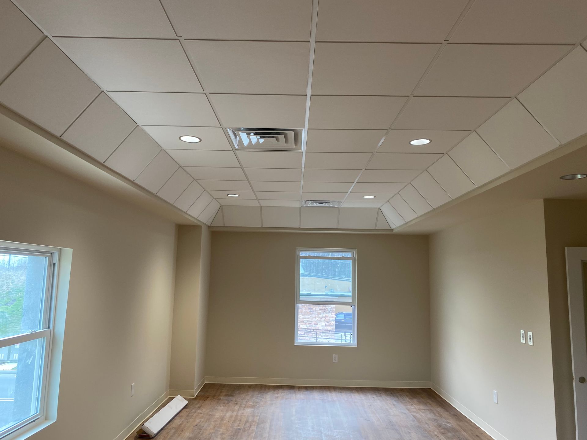 Empty room with drop ceiling, recessed lighting, and a window. Light beige walls, hardwood floor.