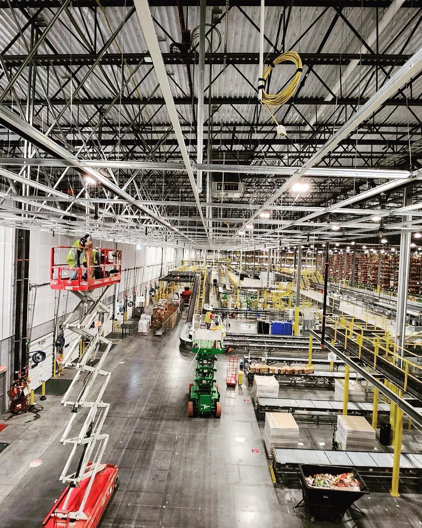 Workers on lifts in a large industrial warehouse; equipment, conveyors, and storage racks.