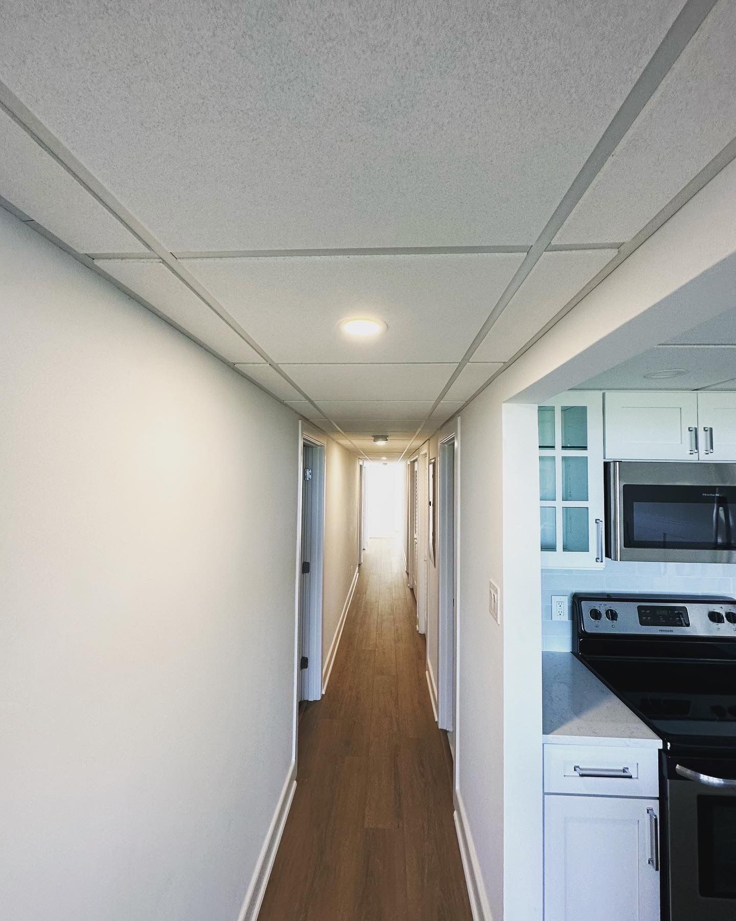 Hallway with white walls, ceiling tiles, and a kitchen visible on the right.