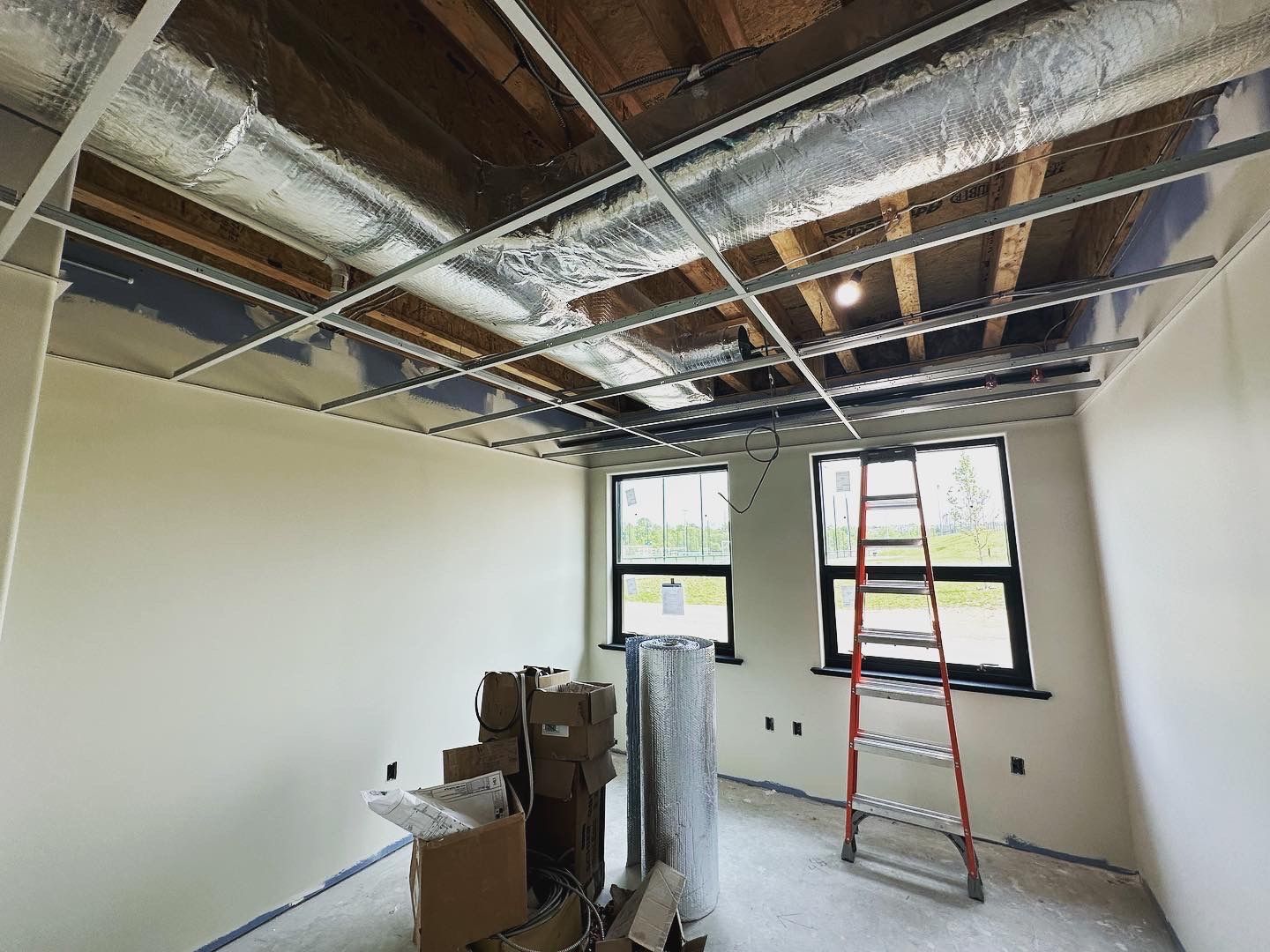Room under construction with exposed ceiling supports, insulation, ladder, windows, and materials.