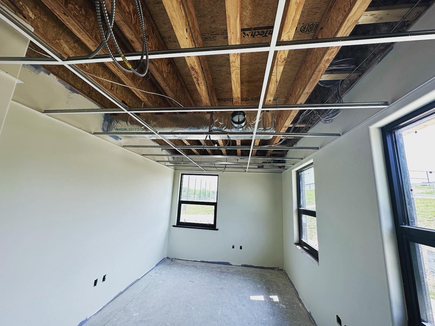 Room under construction; exposed ceiling beams and metal grid, unfinished walls, windows.