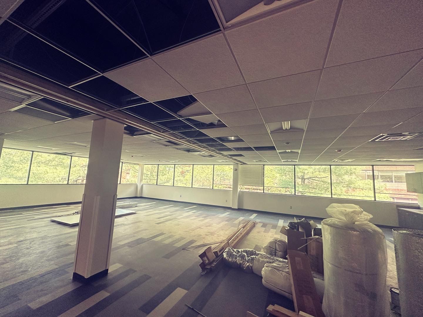 An empty office space under construction, missing ceiling tiles, large windows, and carpeted floor.