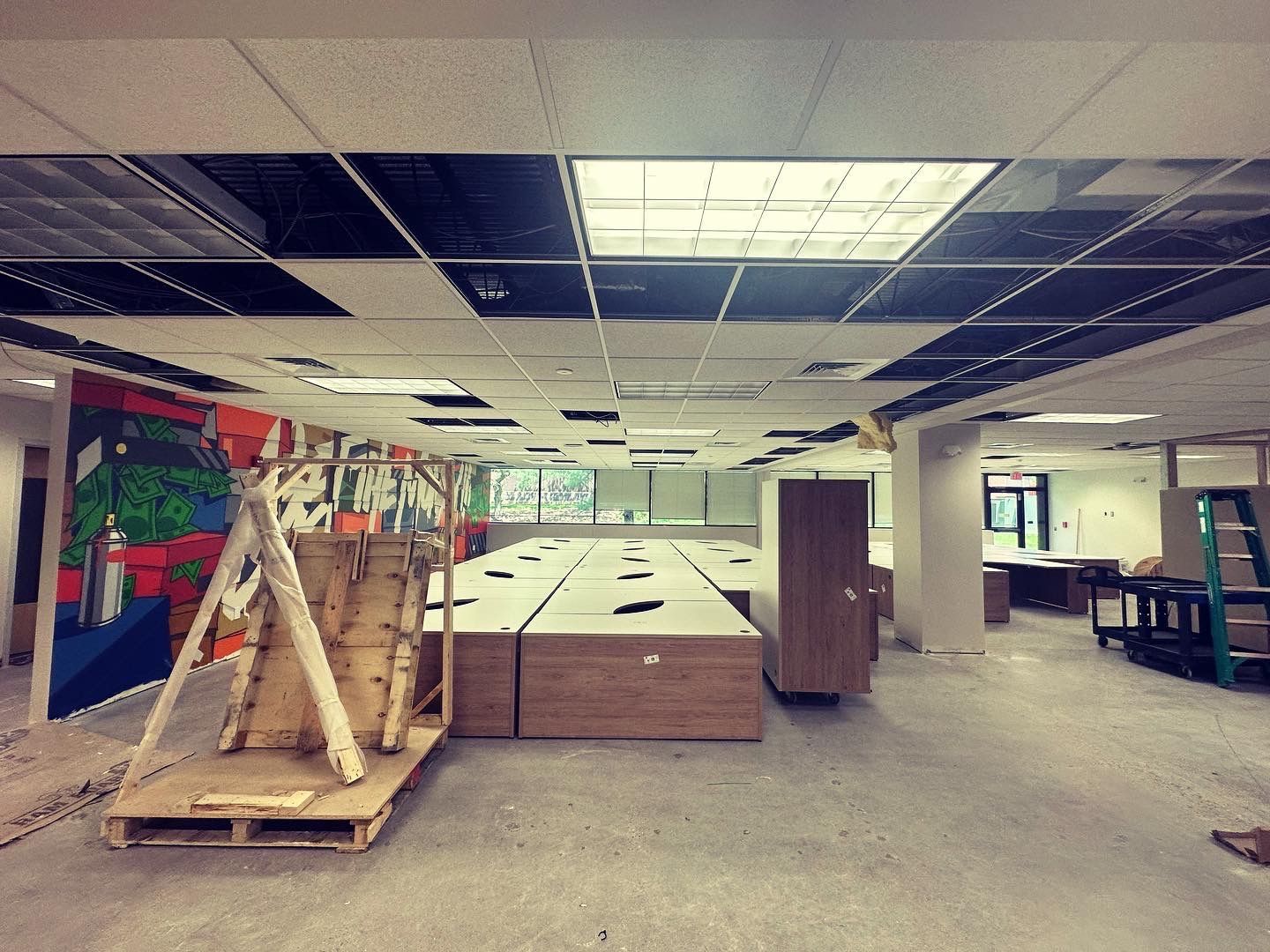 Interior of a workspace under renovation. Ceiling tiles are removed, unfinished flooring, and wood pallets are present.