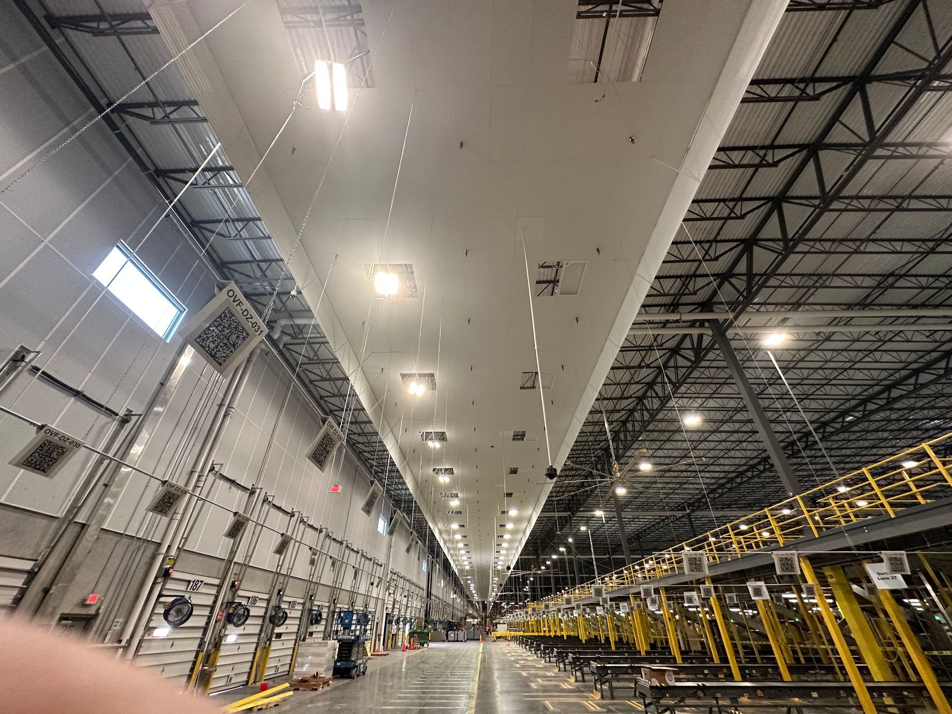 Warehouse interior with long conveyor belt and high ceilings. Rows of workstations on right, loading docks on left.