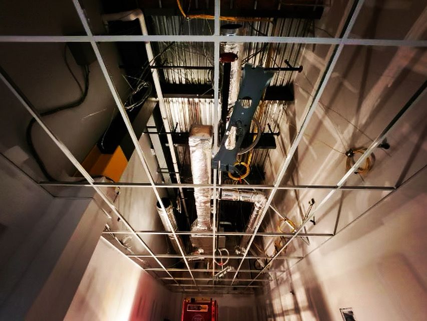 Overhead view of a construction site ceiling grid with exposed pipes, wires, and ductwork.