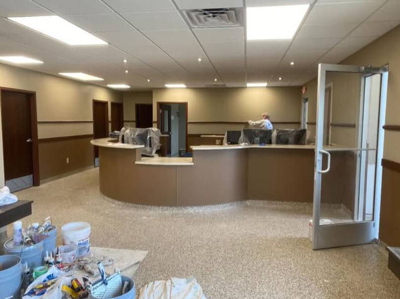 Interior of a building with a curved reception desk. Beige walls, speckled floor, and a partially open door are visible.