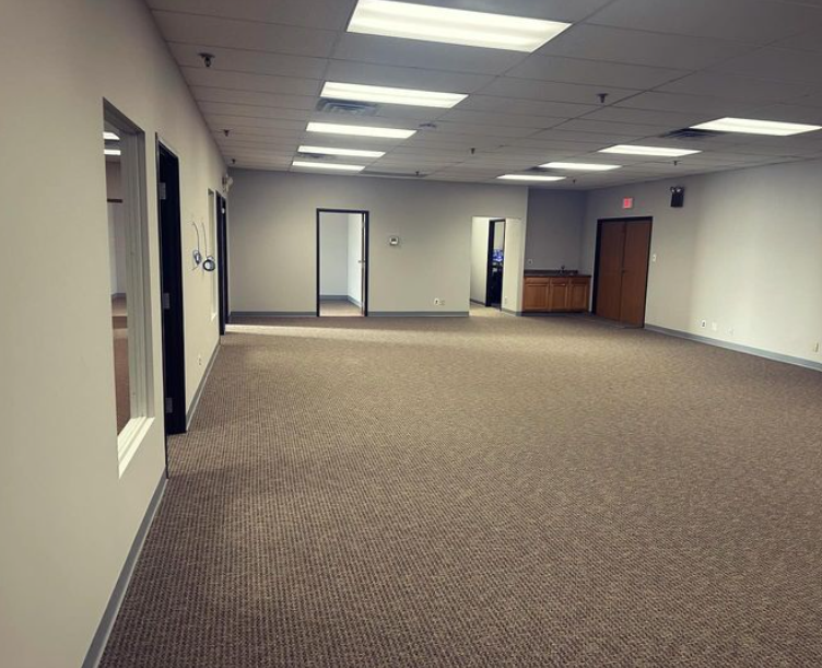 Empty office space with brown carpet, white walls, and overhead lights. Multiple doorways are visible.