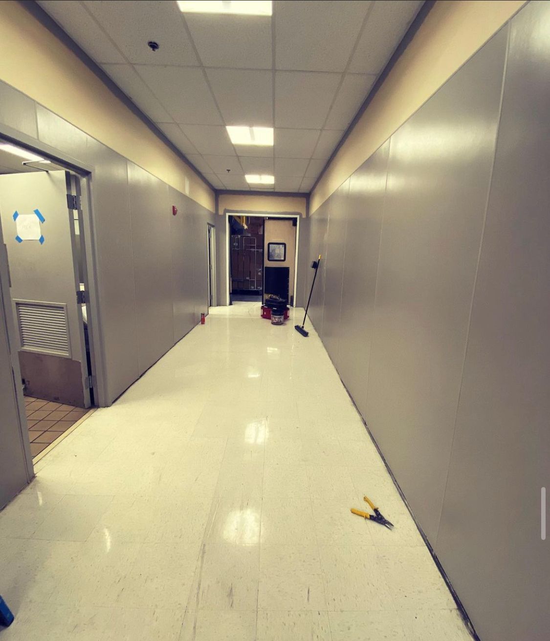 Long hallway with light-colored flooring, gray walls, and open doorways. Tools and equipment sit in the center.