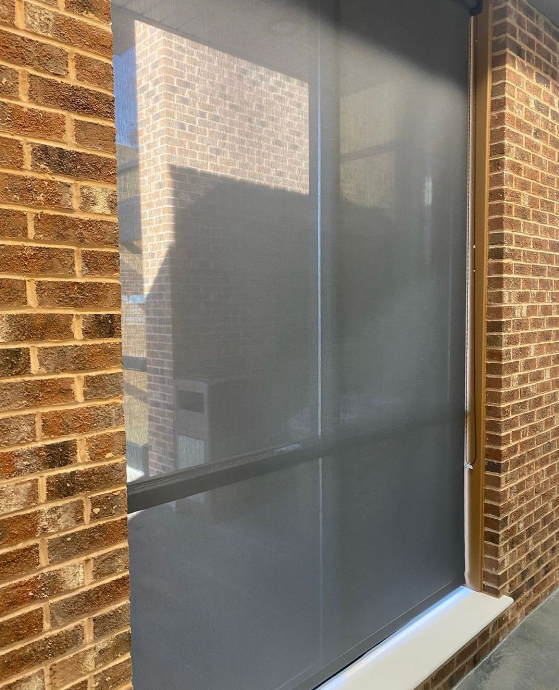 Dark gray roller shade on a brick wall, partially obscuring the view outside.