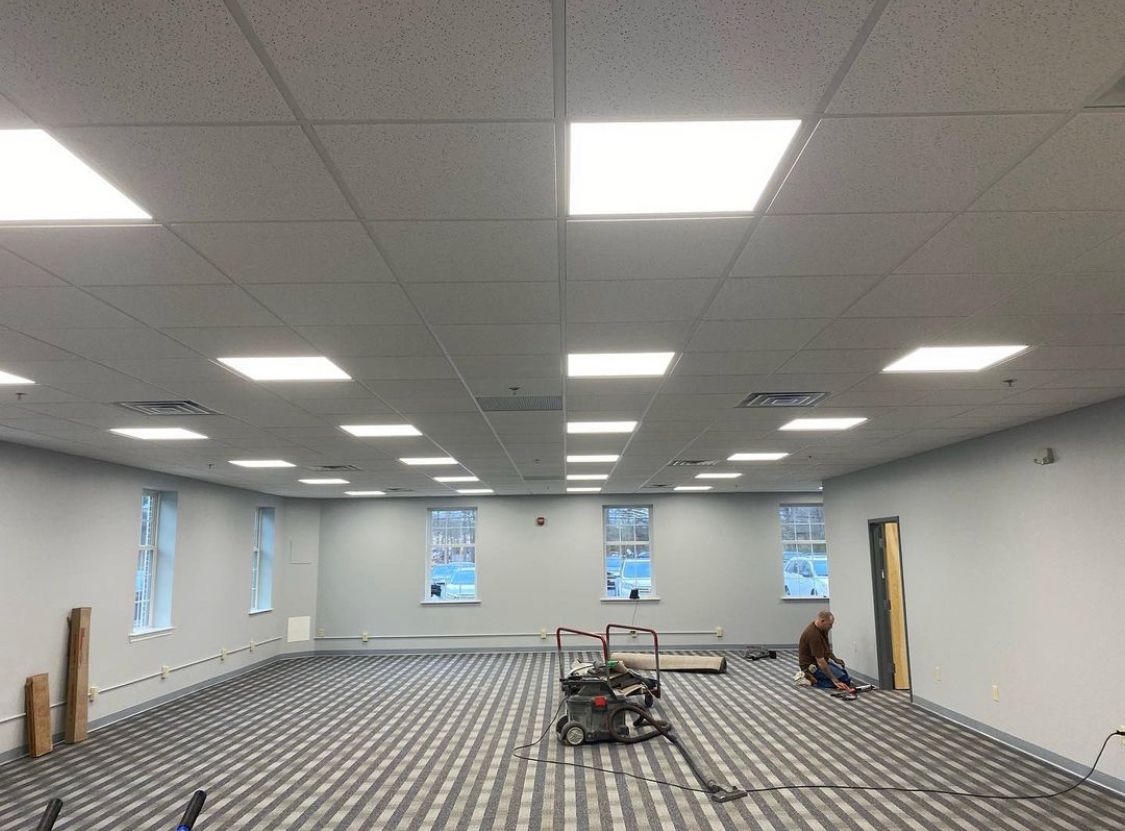 Interior view of a room under construction with new flooring. Ceiling lights are on, worker in distance.