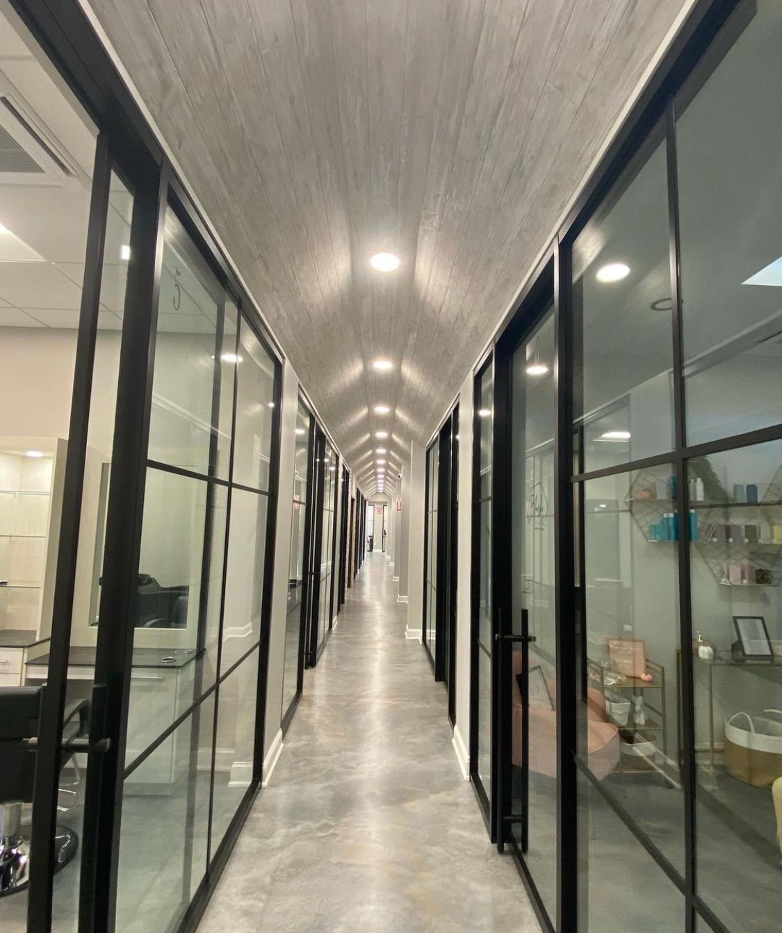 Long hallway with glass-walled offices. Gray floors, black frames, and a light-colored wood-grain ceiling.