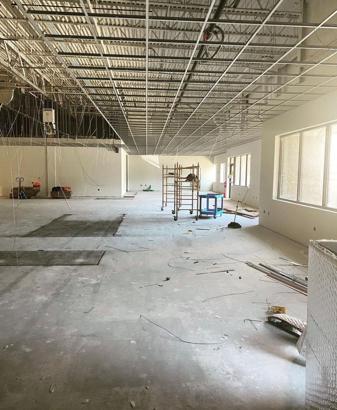 Construction site: unfinished interior with concrete floor, exposed ceiling grid, drywall, ladder, windows, and debris.