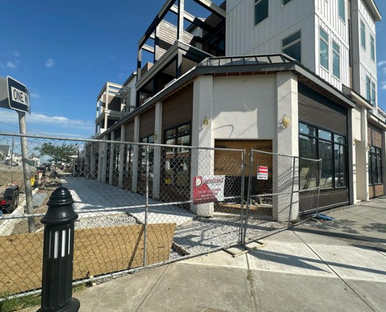Building under construction, fenced off. Sidewalk and street visible. 