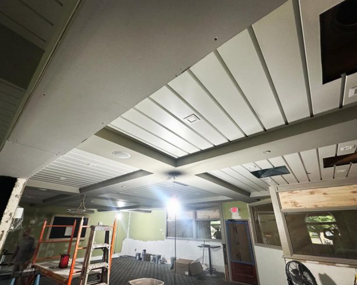 Interior ceiling with angled white panels, overhead lights, and a partially finished construction area.