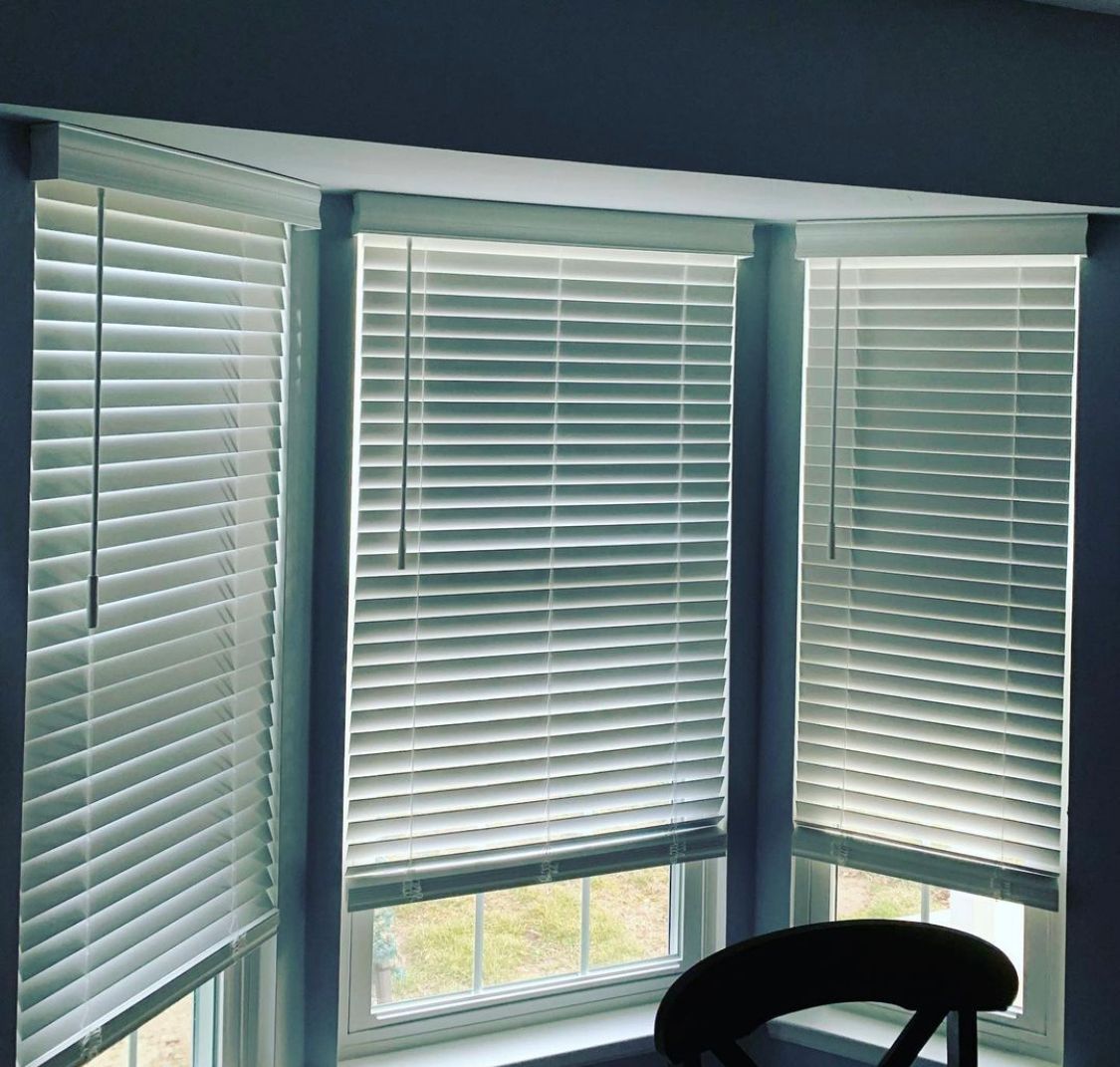 Three white window blinds closed in a bay window, with a partial view of a chair.