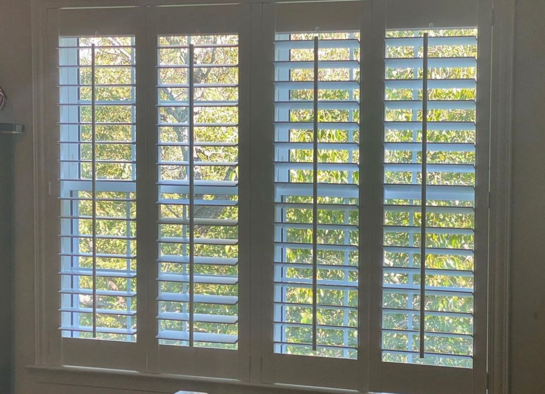 White shutters on a window, slats open to show green foliage outside.