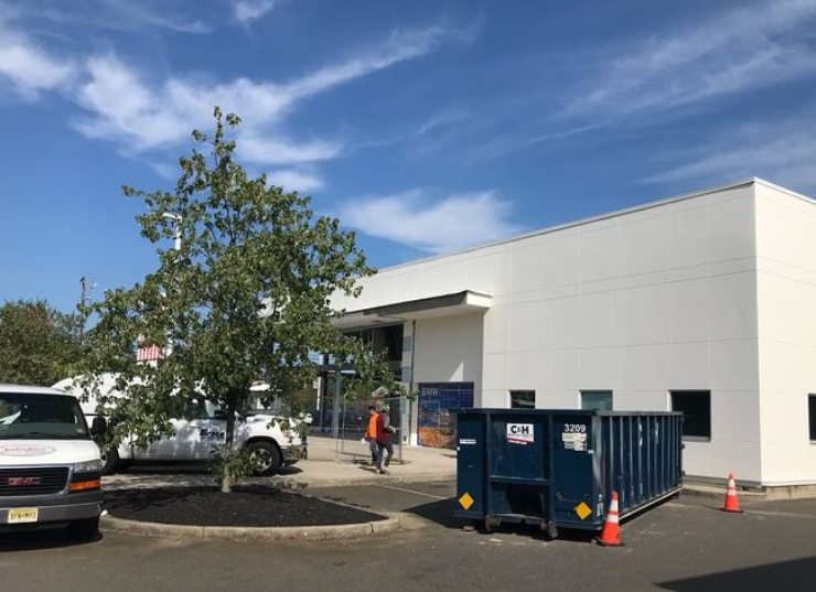 White building with a tree in front. A dumpster and van are in the parking lot. A person walks near the entrance.
