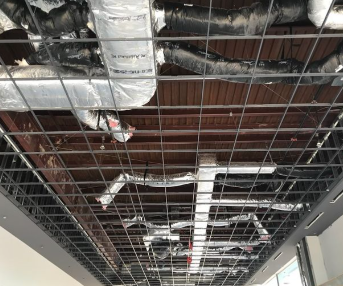 View from below of a metal grid ceiling with exposed ducts and piping against a wood ceiling.
