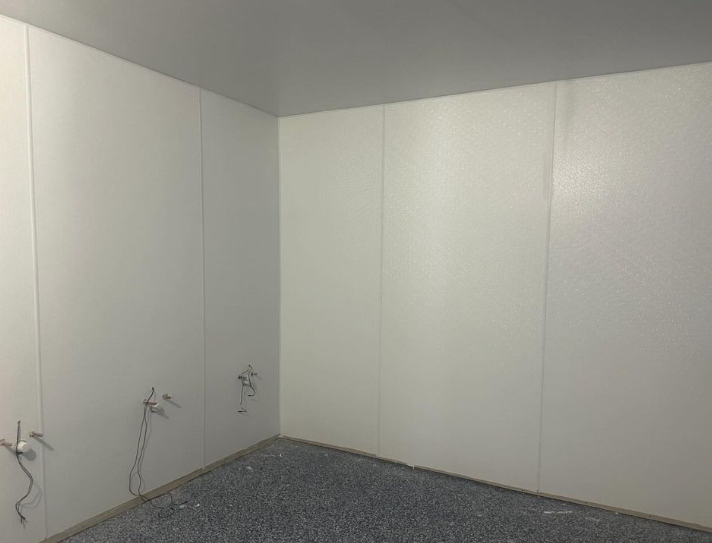 Empty white-paneled room with gray floor and electrical wires.