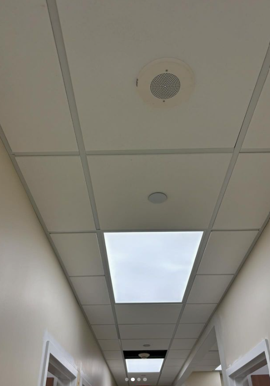 White hallway ceiling with a grid of tiles, recessed lights, and a speaker.