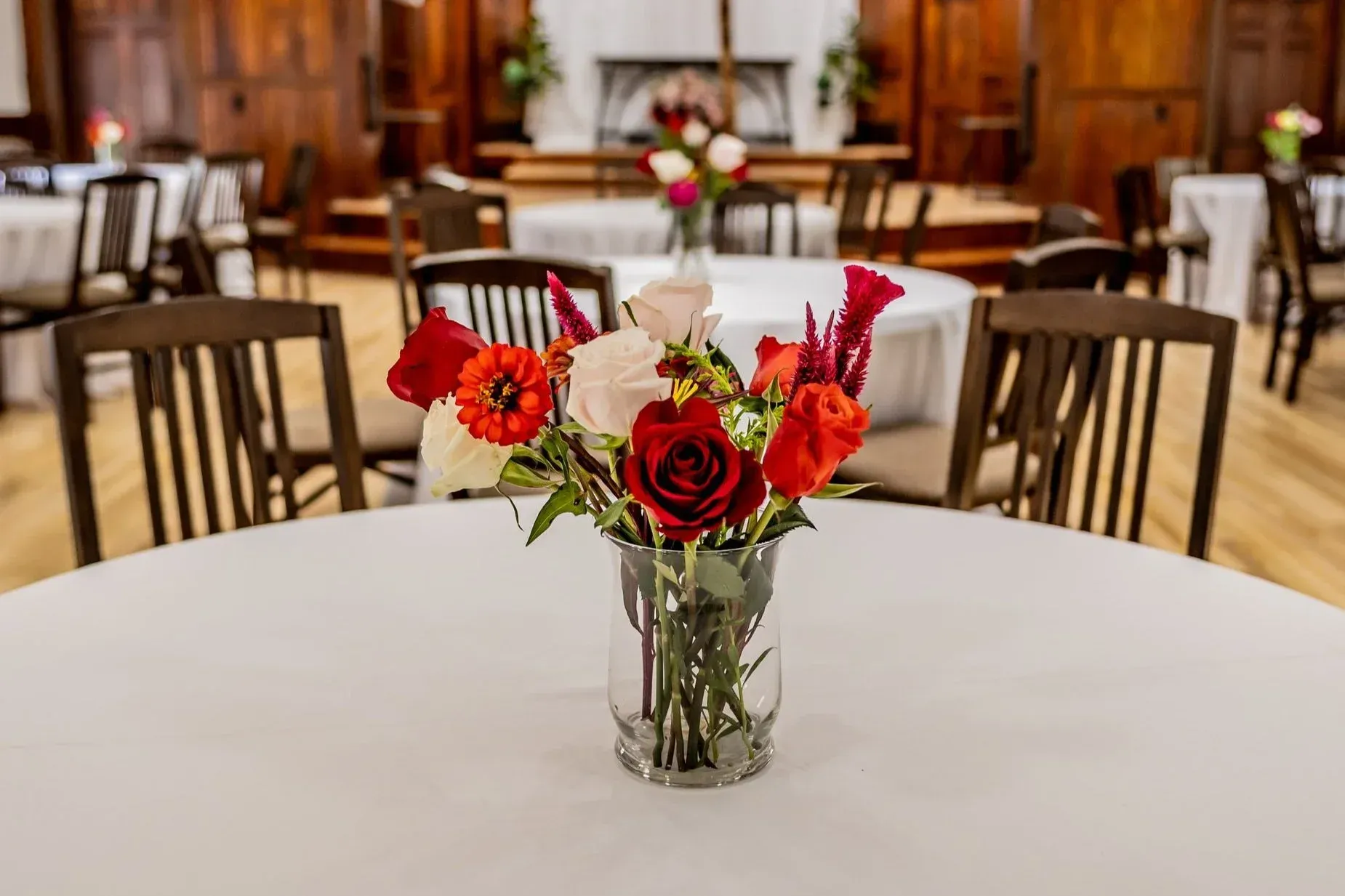 A bouquet of red and white flowers in a glass vase sits on a round, white-clothed table in a wood-paneled banquet hall.