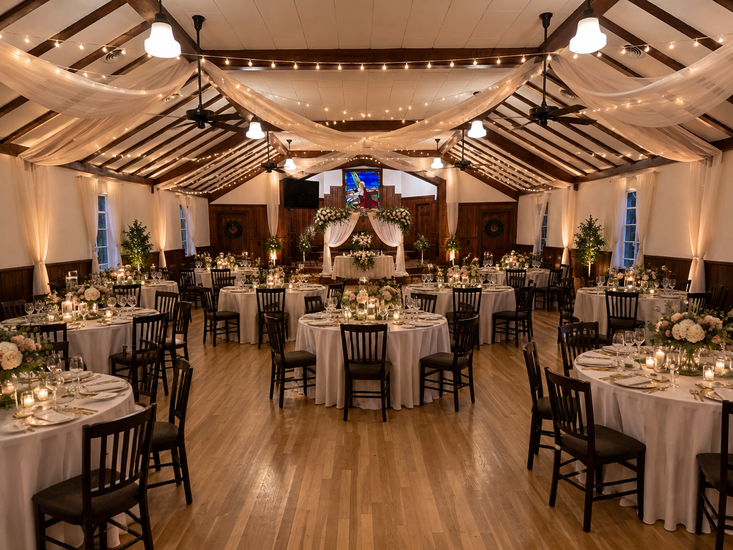 A rustic event hall with wood-paneled walls, a vaulted ceiling, round tables with white cloths, and a stage with art.