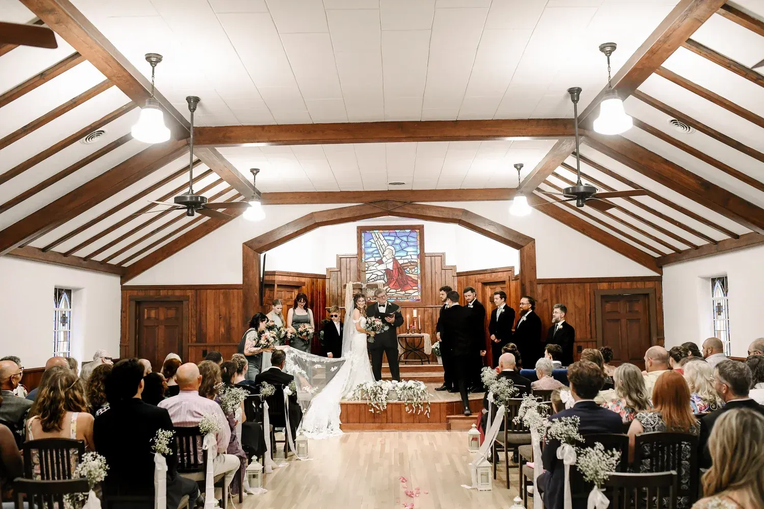 A wedding ceremony takes place in a rustic chapel with wood-paneled walls and exposed beams as guests sit in rows. A wedding ceremony takes place in a rustic chapel with wood-paneled walls and exposed beams as guests sit in rows.