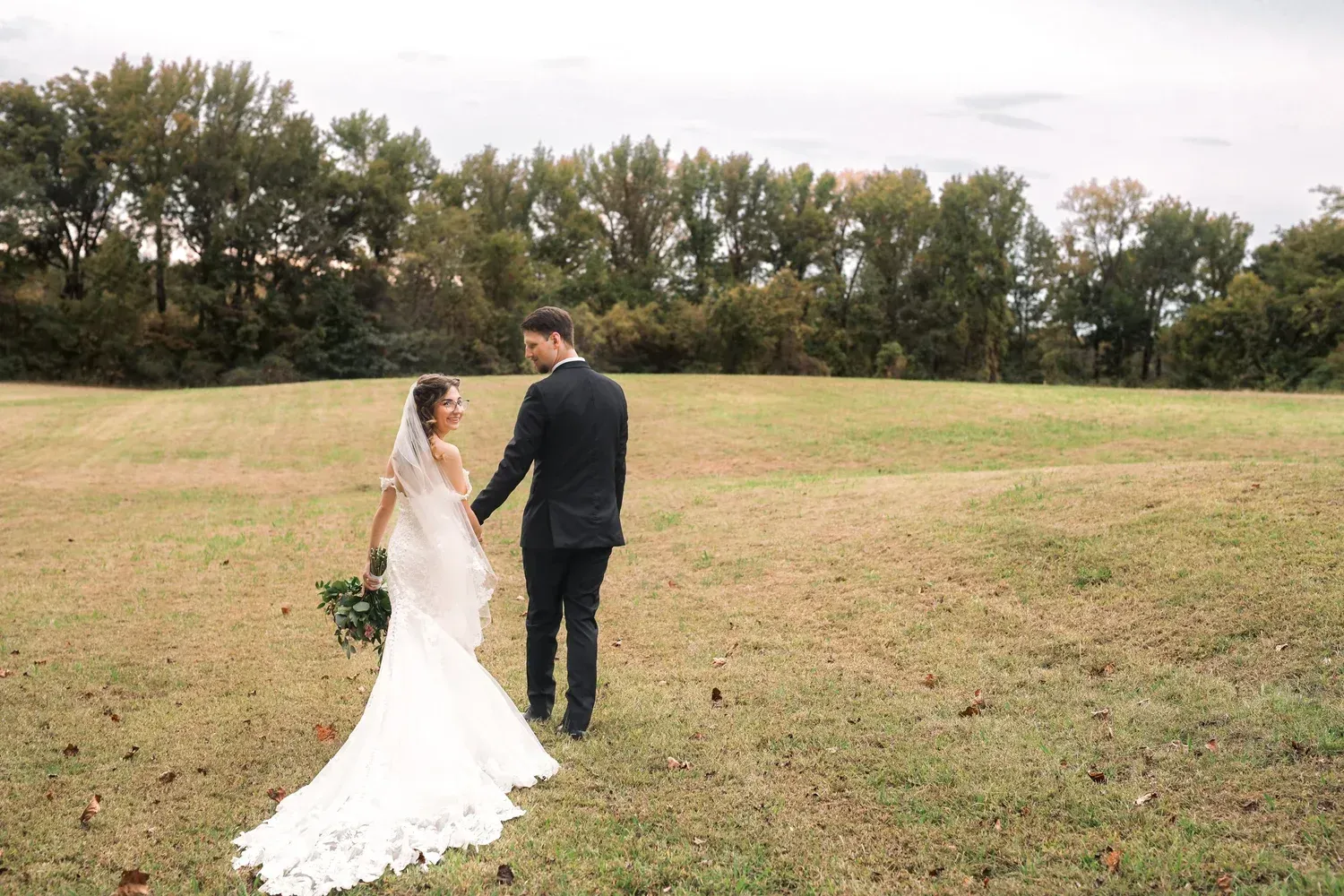 A couple in wedding attire walks hand-in-hand across a grassy field toward a line of trees.