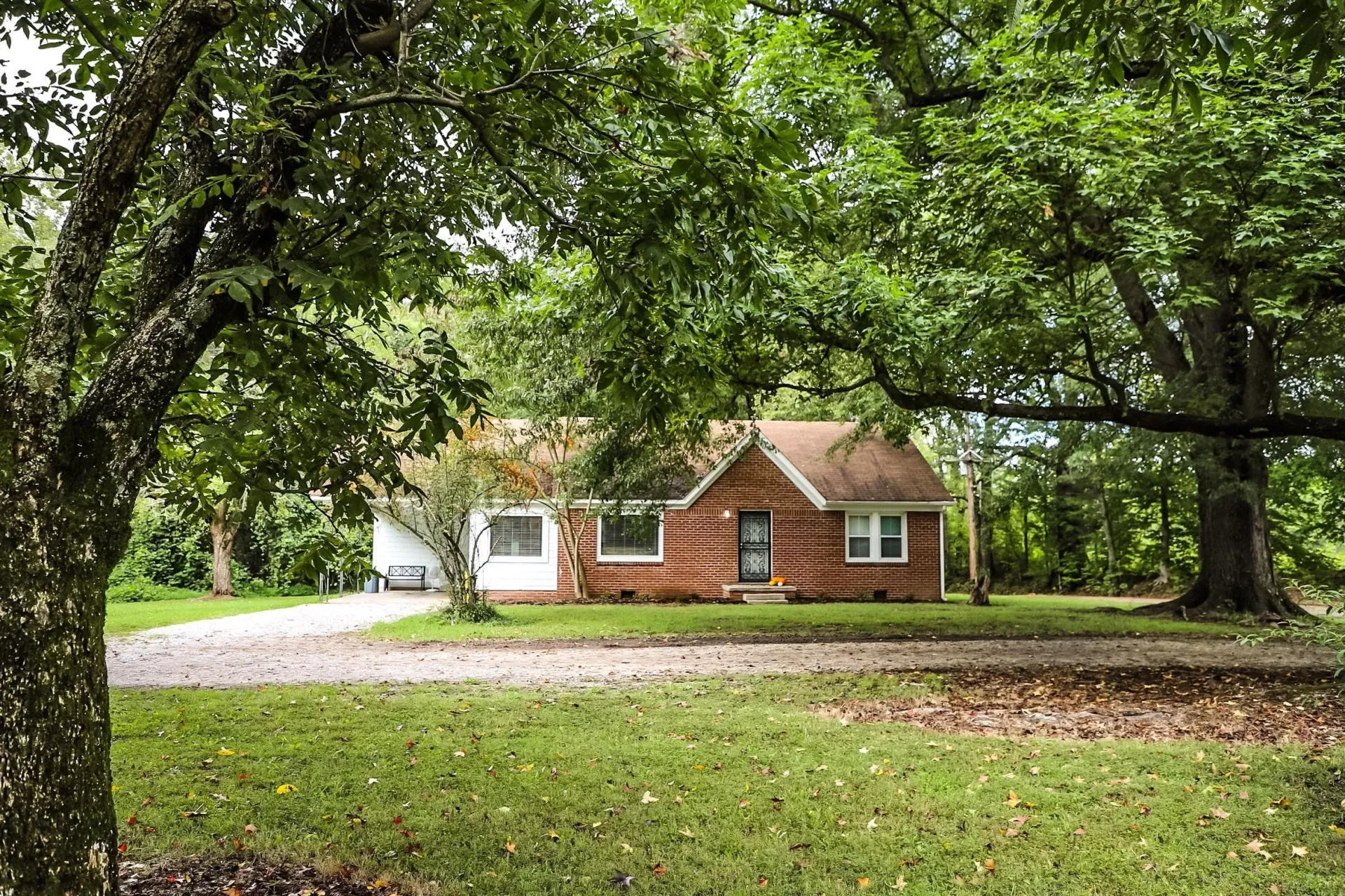 A one-story brick house with a brown roof sits behind large, leafy trees on a grassy, sun-dappled lawn.