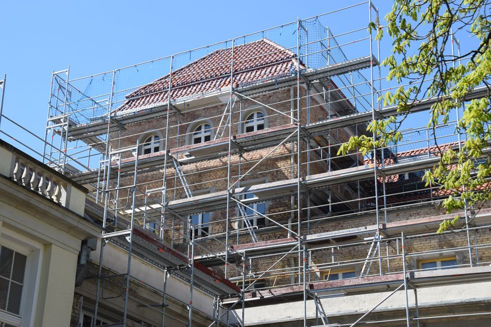 Building under construction with scaffolding around the upper levels; red-tiled roof, windows, clear sky.