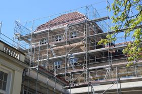 Building under construction with scaffolding around the upper levels; red-tiled roof, windows, clear sky.