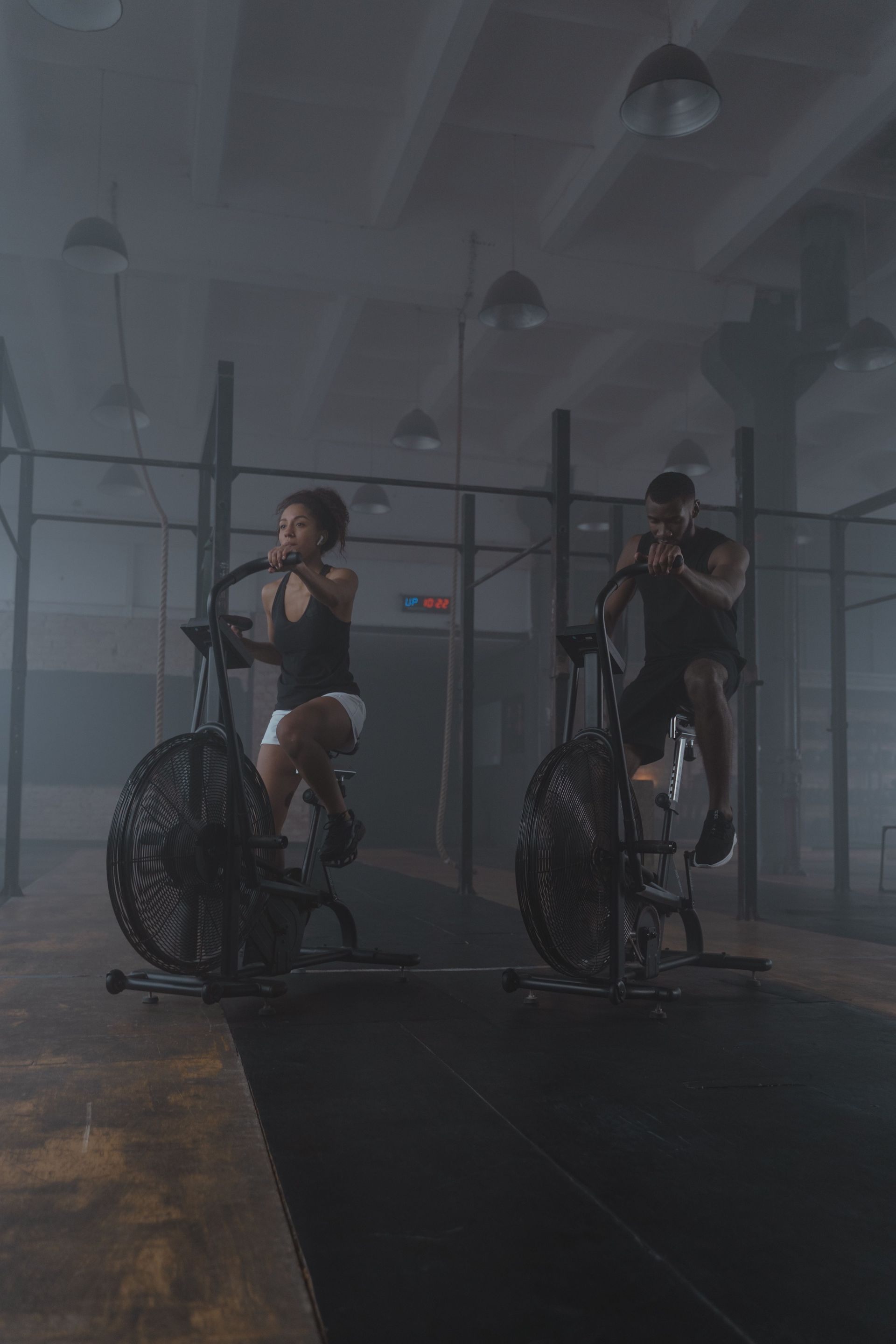 Two cyclists exercising on stationary bikes in a dim gym with industrial lighting