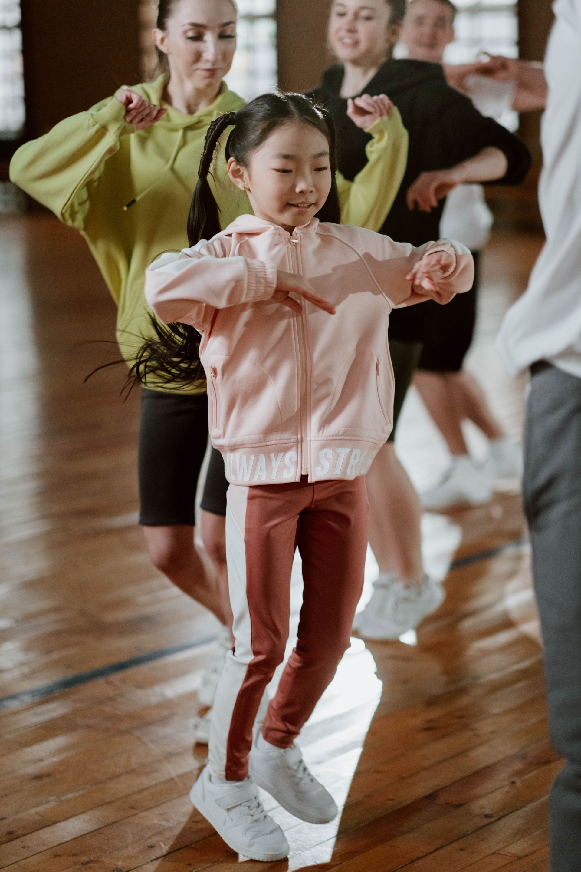 Dancers practicing in a studio, with a child in pink leading the pose in the foreground.