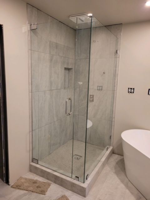 Glass shower enclosure in a bathroom with light gray walls and tile, and a white bathtub.