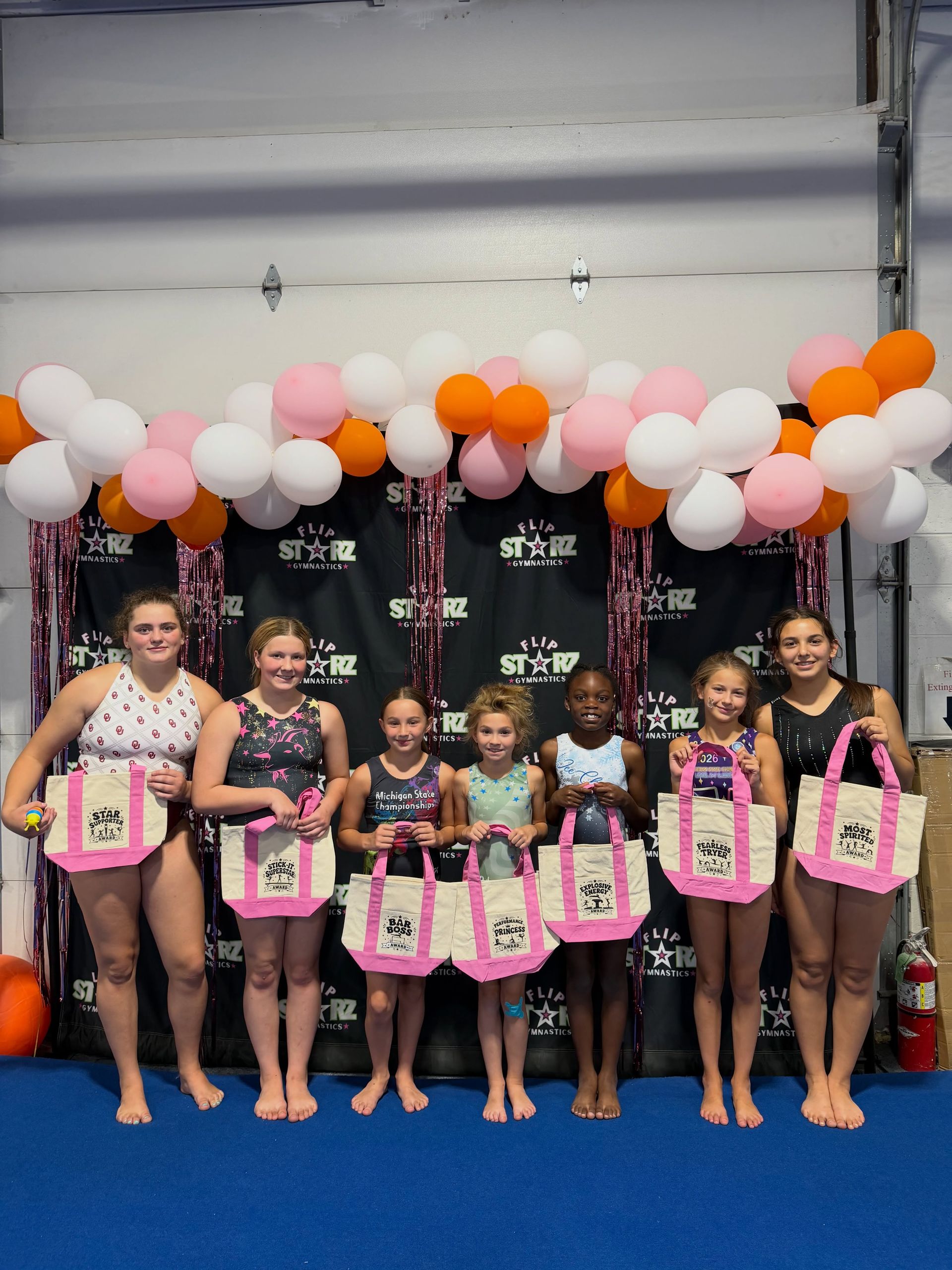 Girls holding pink framed awards under balloon arch on blue mat at a gym event