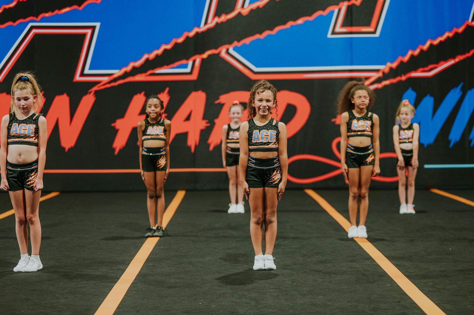 Five cheerleaders in black and gold uniforms stand on a mat in front of a large event banner.