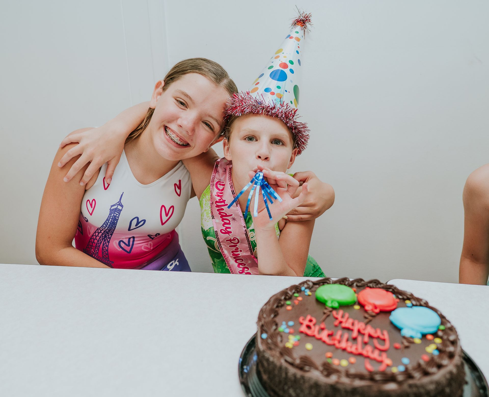 Two children in party hats smile at a birthday table with a chocolate cake in front.