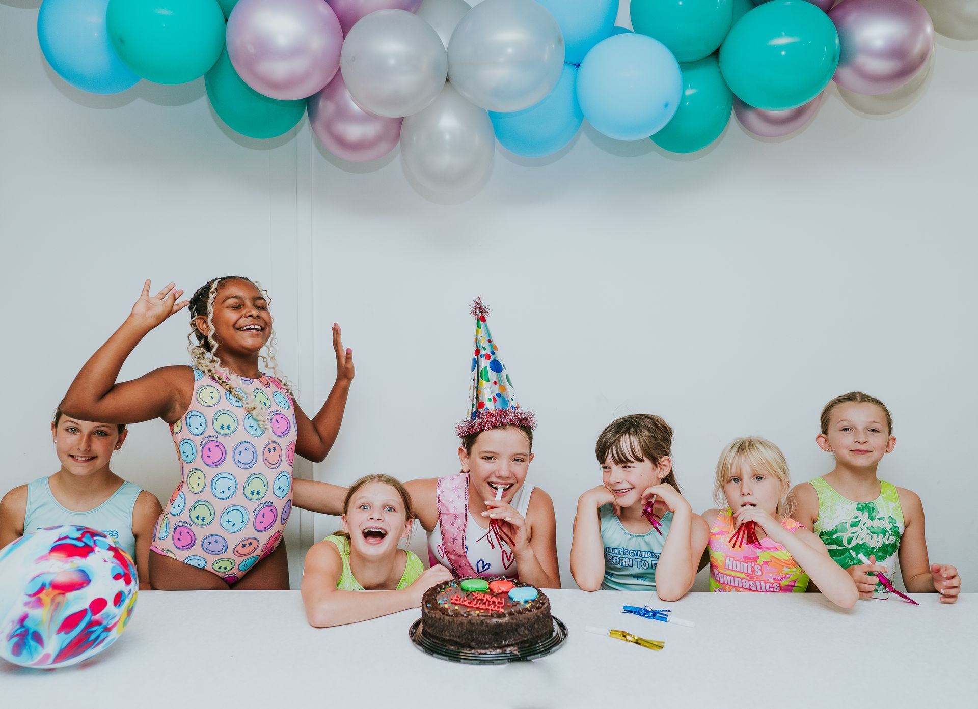 Children at a birthday party, sitting by a cake with balloons overhead and a decorated table