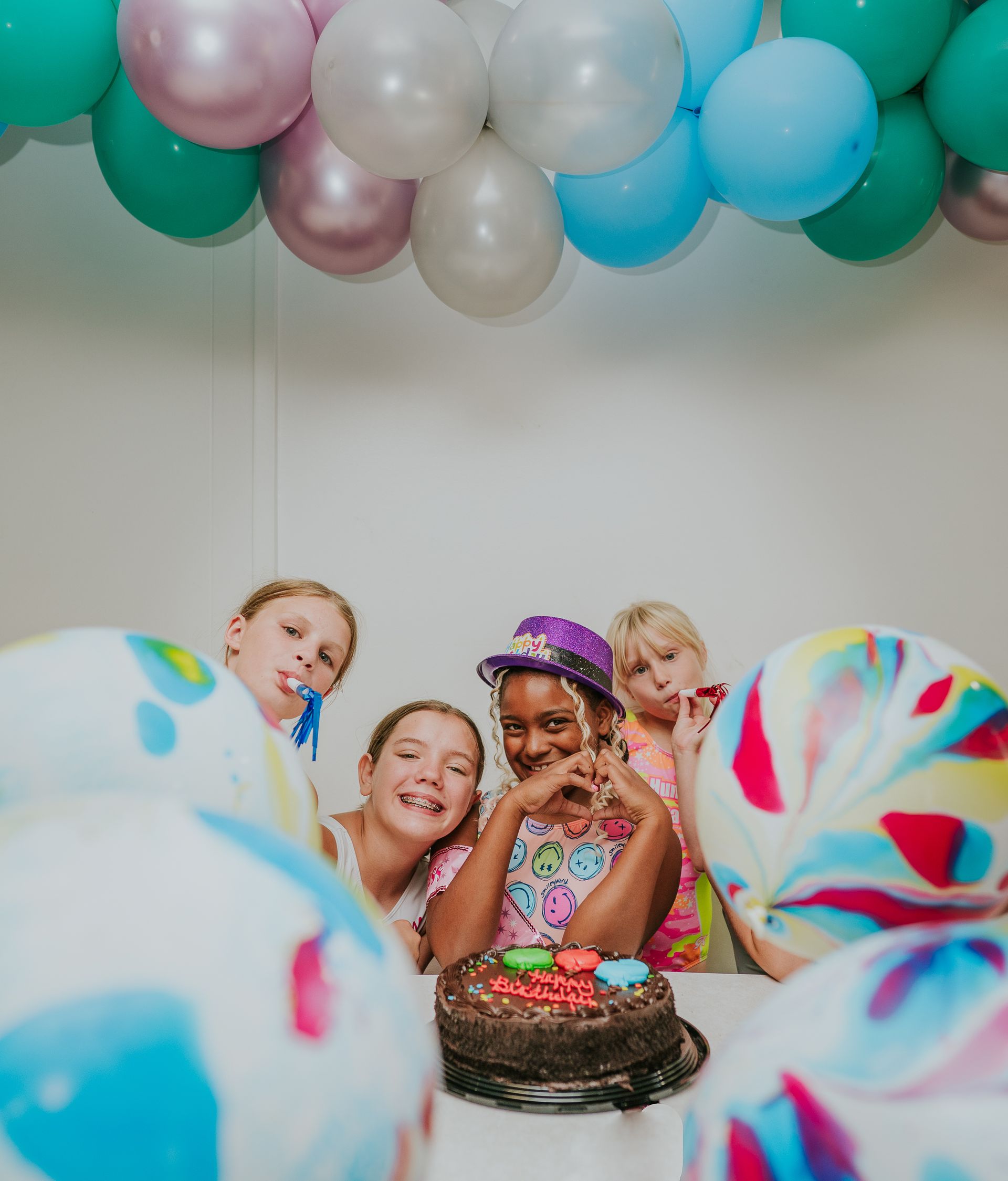 Children gathered around a birthday cake beneath pastel balloons, smiling and celebrating.