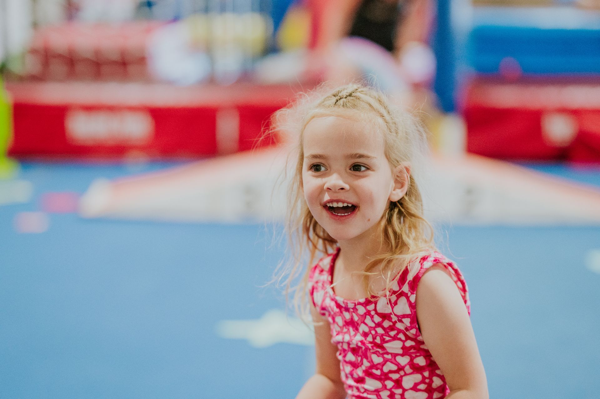 Smiling blonde child in a pink outfit at an indoor gymnastics gym