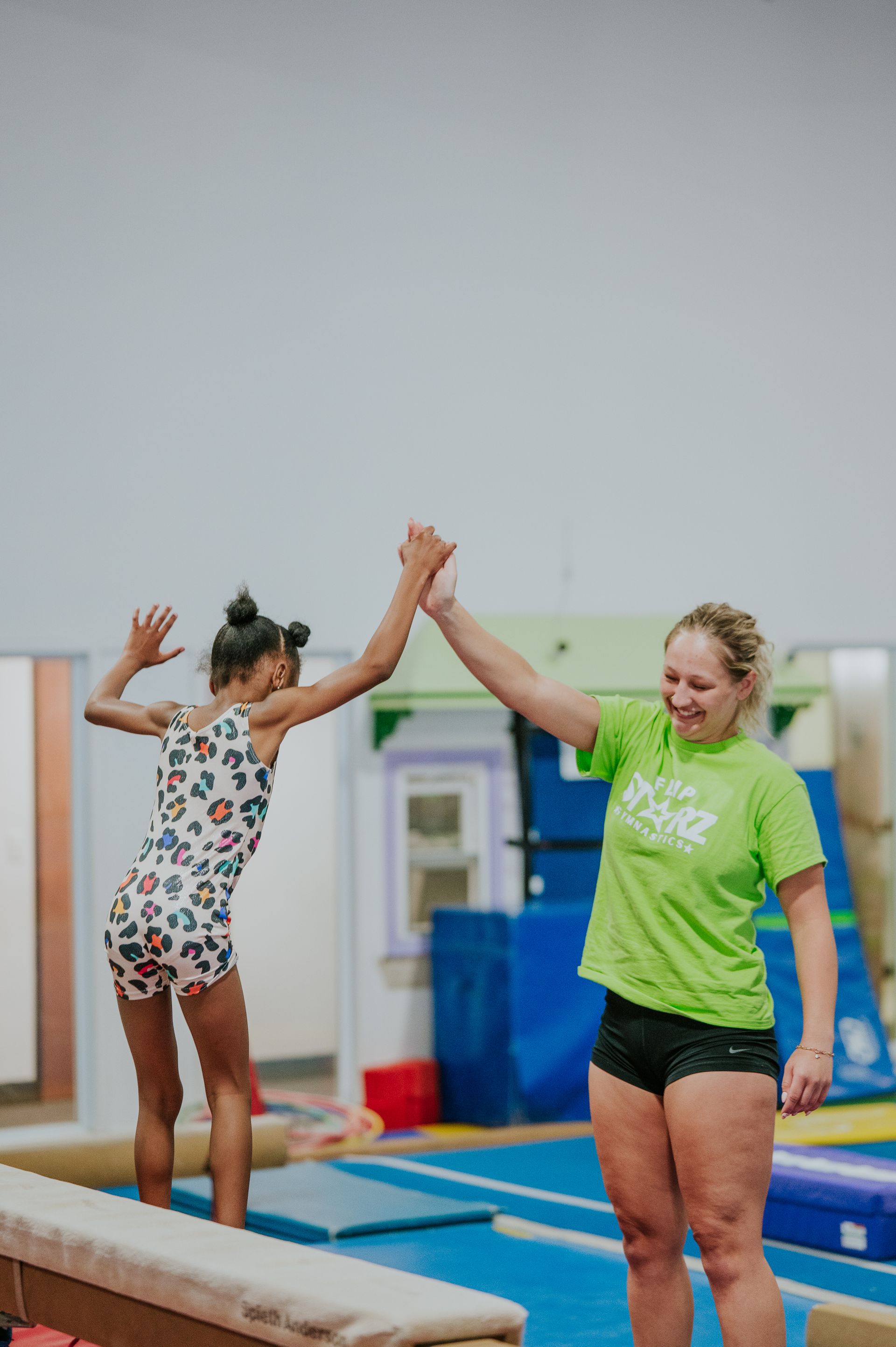 Two gymnasts high-five on a gymnastics floor with balance beams in the background.