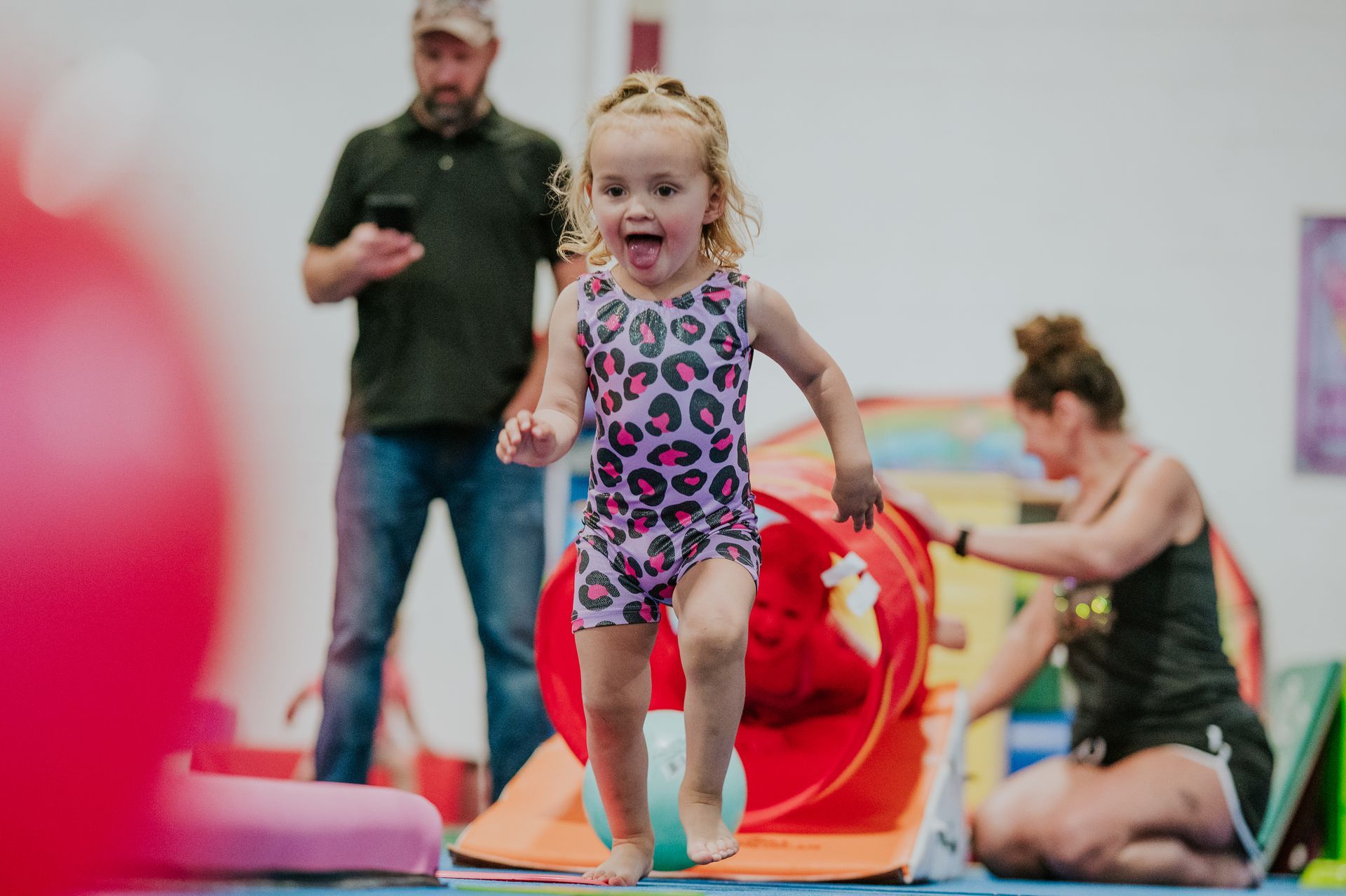 Toddler running indoors on a colorful play mat, with adults in the background supervising nearby.