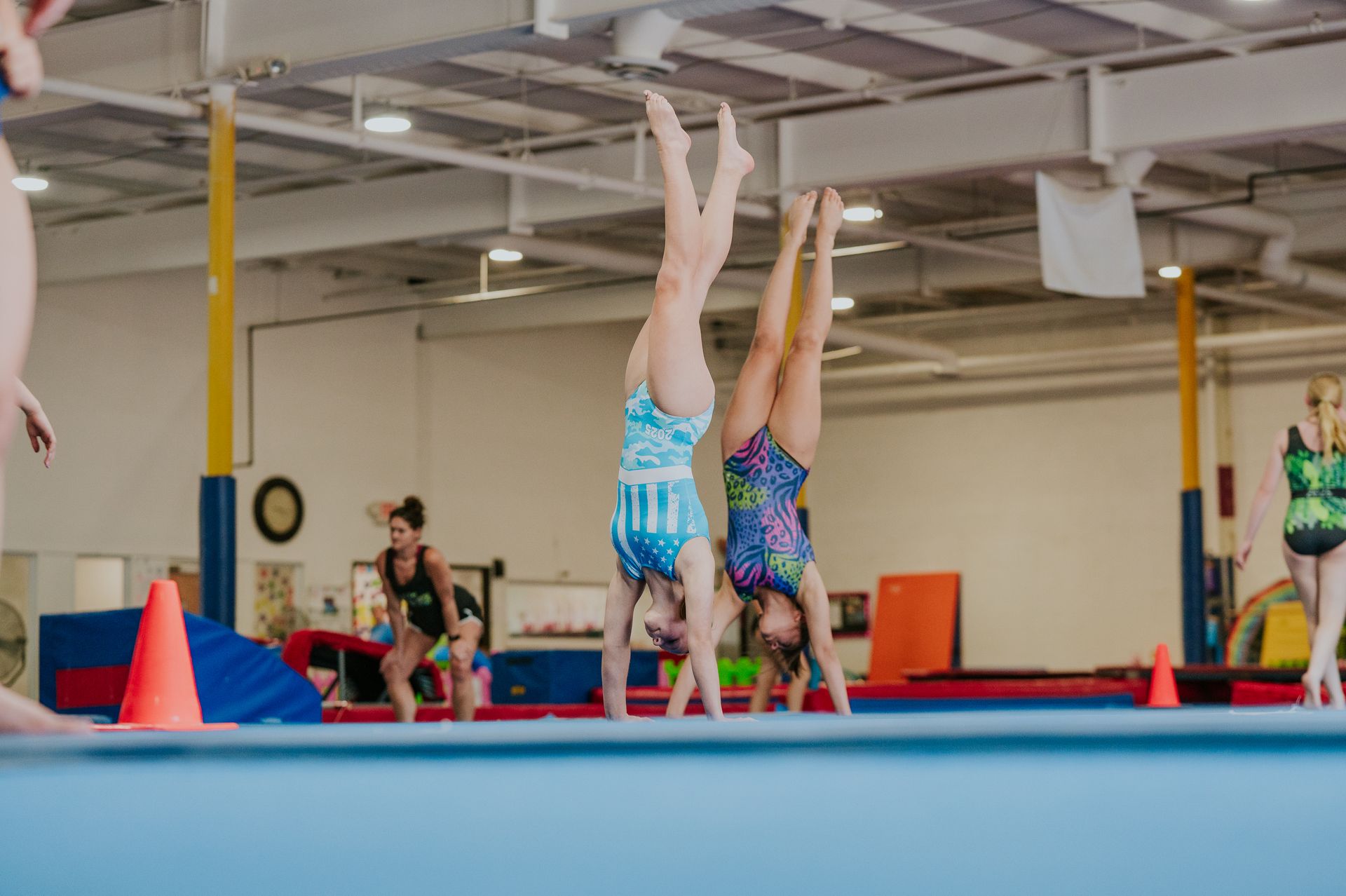 Gymnastics practice in a gym, with two athletes handstanding on a blue mat near orange cones.