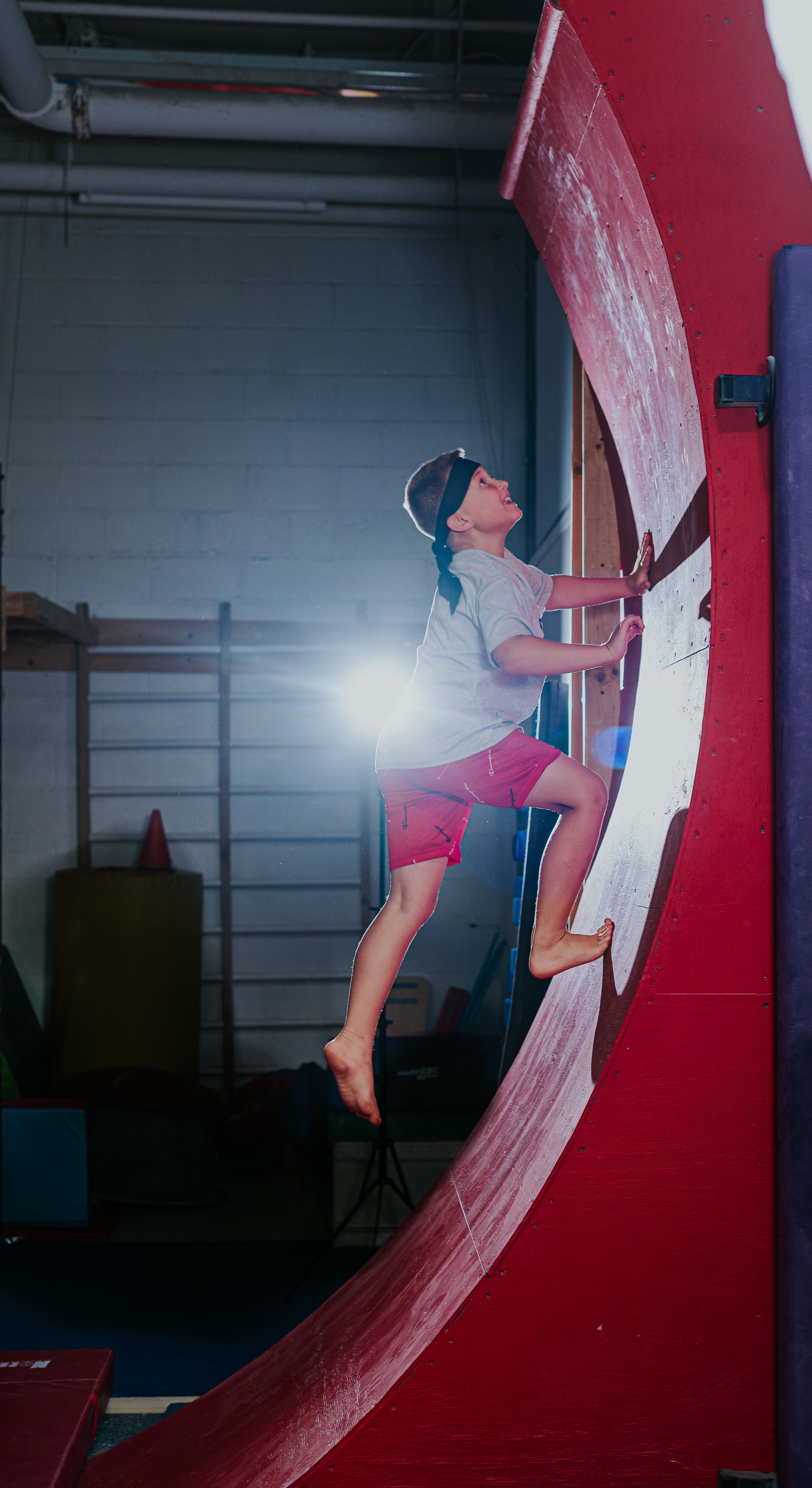 Person climbing a steep red indoor rock wall, reaching upward with a bright light behind them.