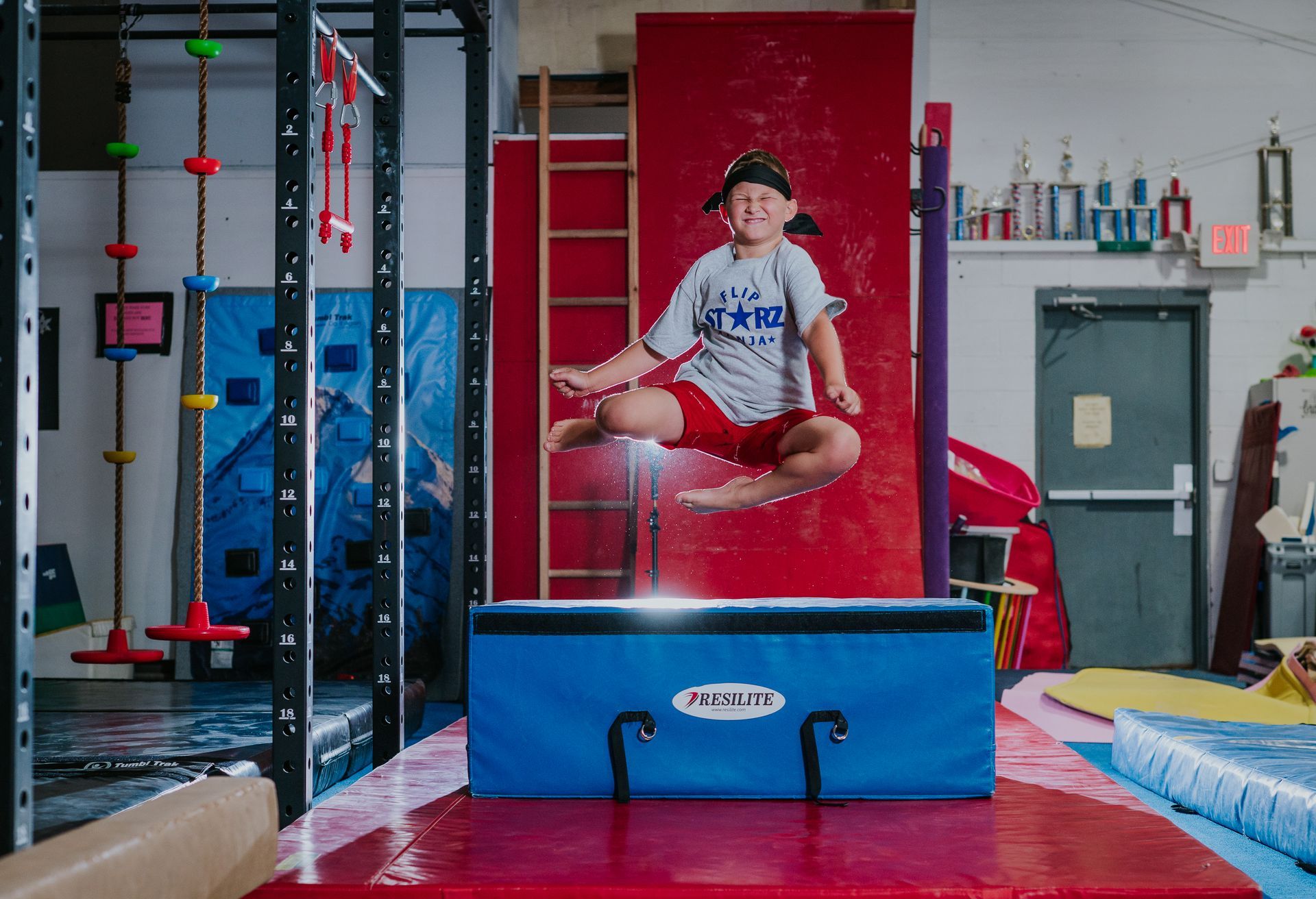 Child midair on a red gymnastics mat in an indoor gym, near climbing equipment and a blue crash mat