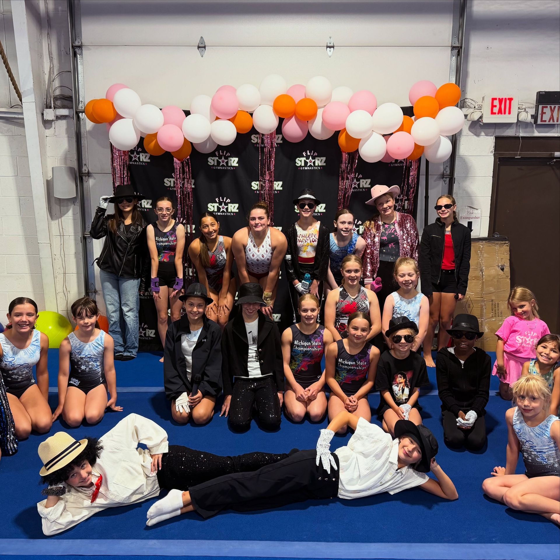 Large group posing in dance costumes on a blue floor beneath a pink, white, and orange balloon arch