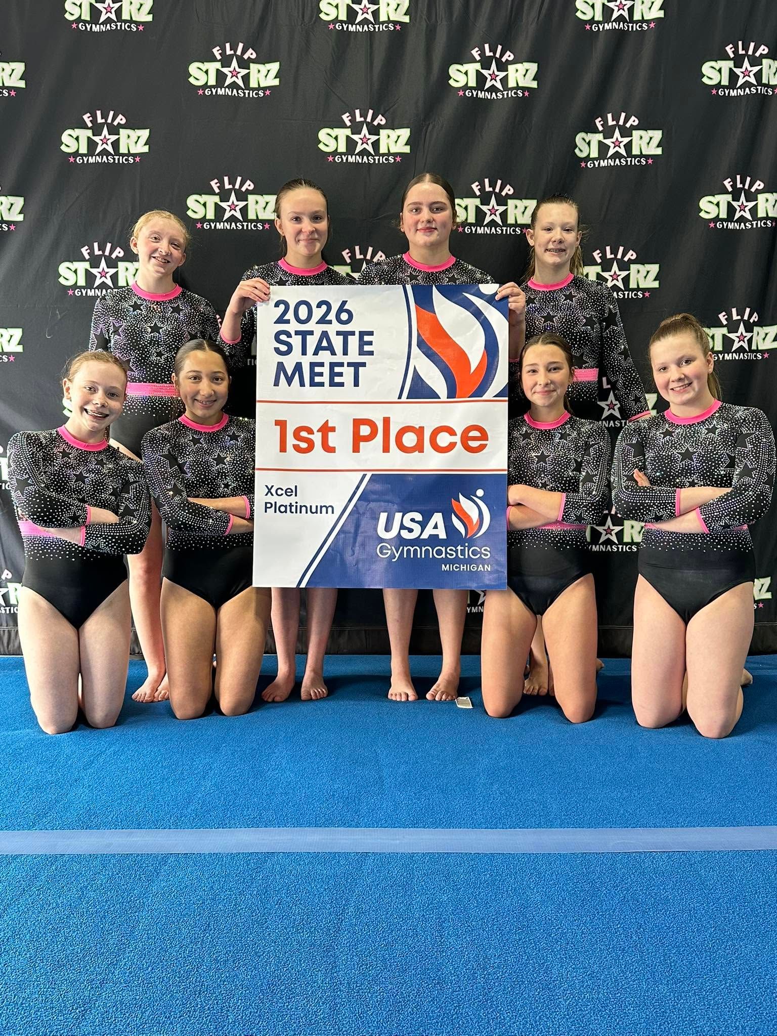 Cheer team posing with 2026 State Meet 1st Place sign on blue mat, standing before sponsor backdrop